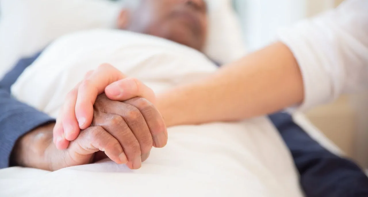 Mano de una persona cuidadora sosteniendo la mano de una persona mayor en la cama
